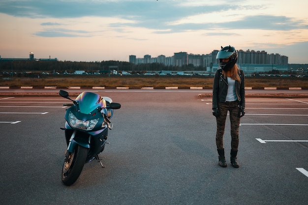 Retrato de cuerpo entero de una elegante joven mujer caucásica con jeans caqui, chaqueta de cuero negro y casco protector de pie en el estacionamiento y mirando la moto azul estacionada junto a ella