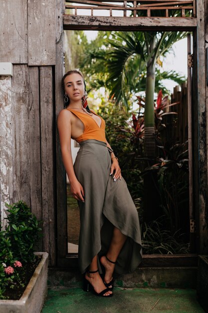 Retrato de cuerpo entero de elegante mujer elegante con accesorios en la cabeza y falda larga y tacones posando en un parque exótico