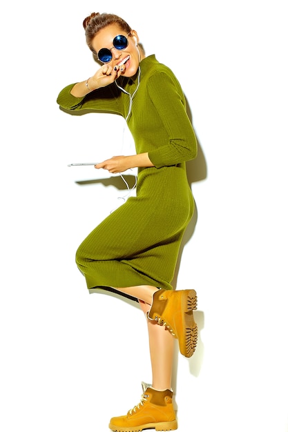 Retrato de cuerpo entero de hermosa feliz linda morena sonriente mujer niña en ropa de verano casual hipster verde aislado en blanco escuchando música en smartphone con auriculares