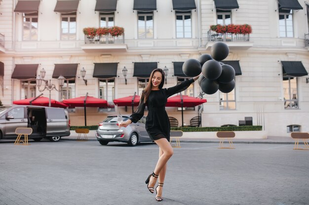 Retrato de cuerpo entero de una hermosa mujer morena en zapatos elegantes bailando con globos de fiesta en la calle