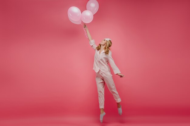 Retrato de cuerpo entero de una hermosa niña despreocupada de pie de puntillas con globos. Foto interior de dama rizada en ropa de dormir rosa y antifaz saltando con una sonrisa.