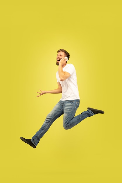 Retrato de cuerpo entero de hombre feliz saltando aislado sobre fondo amarillo. Modelo masculino caucásico en ropa casual. Libertad de elección, inspiración, concepto de emociones humanas. Date prisa, hablando por teléfono.