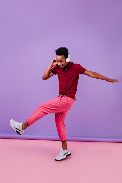 Retrato de cuerpo entero de un hombre inspirado con estilo bailando en auriculares. Filmación en interiores de chico africano emocional bromeando.