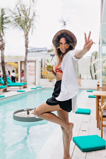 Retrato de cuerpo entero de una impresionante mujer descalza escalofriante junto a la piscina. Retrato al aire libre de adorable mujer morena con sombrero y gafas de sol divirtiéndose en el resort.