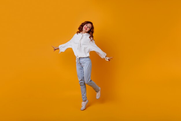 Retrato de cuerpo entero de una increíble chica delgada con cabello pelirrojo. Mujer romántica caucásica bailando en la pared amarilla.