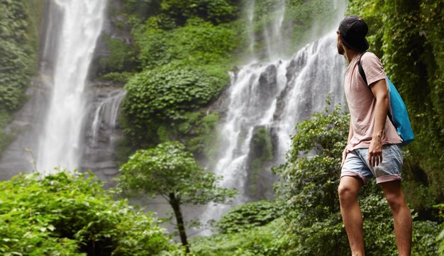 Retrato de cuerpo entero de joven excursionista o aventurero en pantalones cortos de mezclilla y snapback disfrutando de la naturaleza