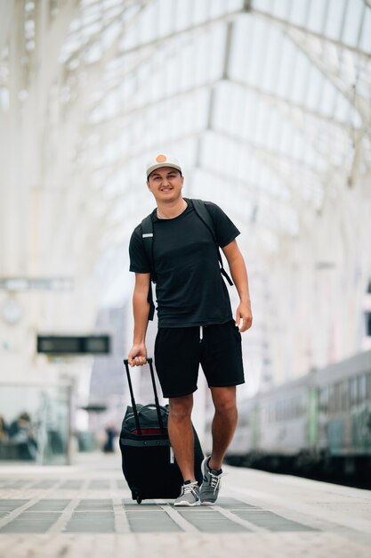 Retrato de cuerpo entero de un joven feliz caminando con maleta en la estación de tren