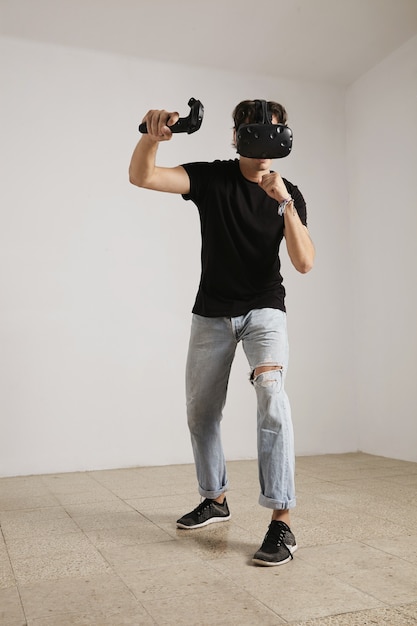 Retrato de cuerpo entero de un joven jugador con gafas de realidad virtual y jeans y una camiseta negra sin etiqueta que juega a un juego en una habitación con paredes blancas y piso de madera clara.