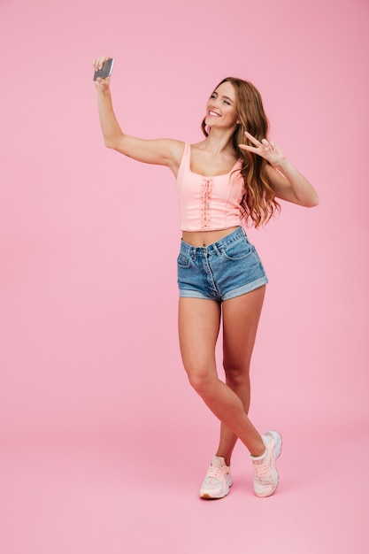 Retrato de cuerpo entero de una joven mujer sonriente de cabeza lectora en ropa de verano hace selfie en teléfono inteligente, mostrando el signo de la paz