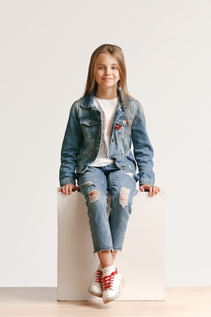 Retrato de cuerpo entero de linda niña adolescente en ropa de jeans con estilo mirando a cámara y sonriendo contra la pared blanca del estudio. Concepto de moda infantil