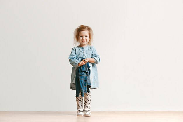 Retrato de cuerpo entero de una linda niña pequeña en ropa de jeans con estilo y sonriente, de pie en blanco. Concepto de moda infantil