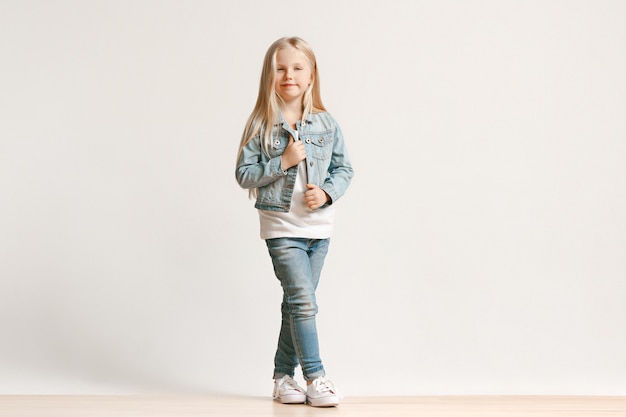 Retrato de cuerpo entero de una linda niña pequeña en ropa de jeans con estilo y sonriente, de pie en blanco. Concepto de moda infantil