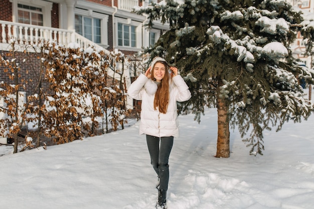 Retrato de cuerpo entero de la moda dama de pelo largo posando al lado de la casa cerca del árbol nevado verde. Foto al aire libre de adorable mujer caucásica en chaqueta pasar el invierno en el patio disfrutando del buen tiempo.