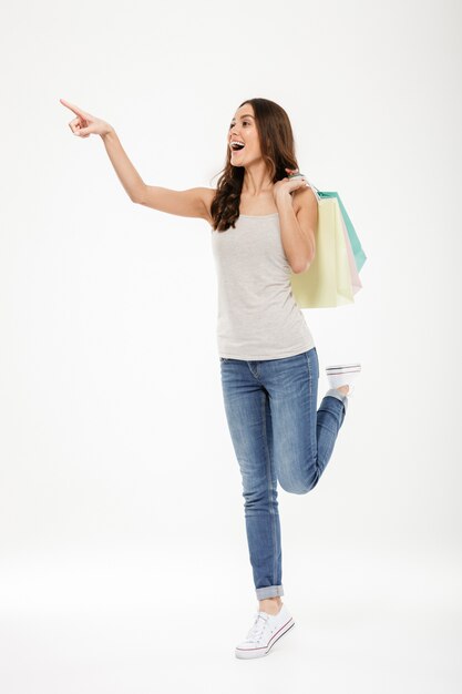 Retrato de cuerpo entero de moda mujer apuntando con el dedo y sosteniendo muchas bolsas de compras, aislado sobre la pared blanca