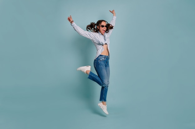 Retrato de cuerpo entero del modelo femenino encantador divertido saltando y divirtiéndose sobre la pared aislada. Niña feliz baila y sonríe sobre fondo azul aislado.