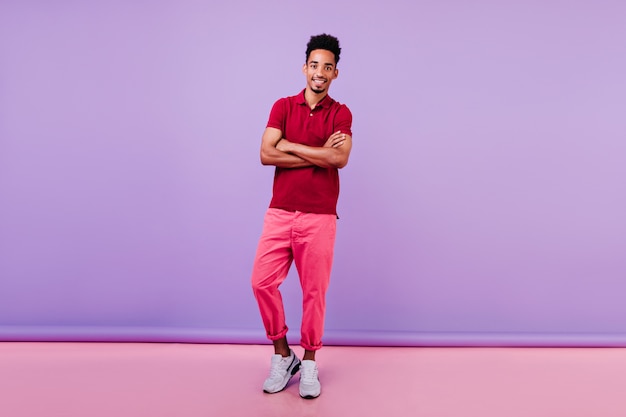 Retrato de cuerpo entero del modelo masculino interesado en pantalón rosa. Hombre joven negro despreocupado de pie con los brazos cruzados.