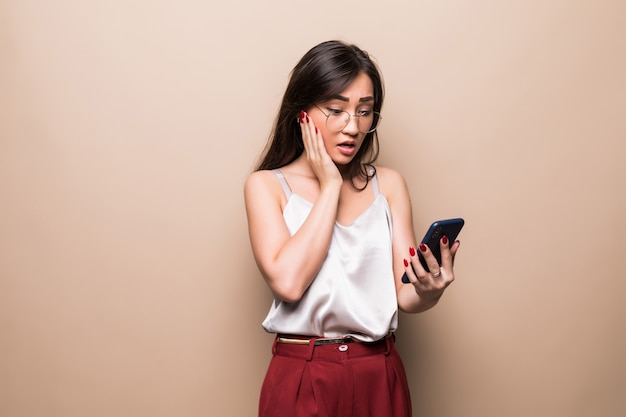 Retrato de cuerpo entero de mujer asiática sorprendida usando teléfono móvil mientras sostiene la taza de café para ir aislado sobre la pared de color beige