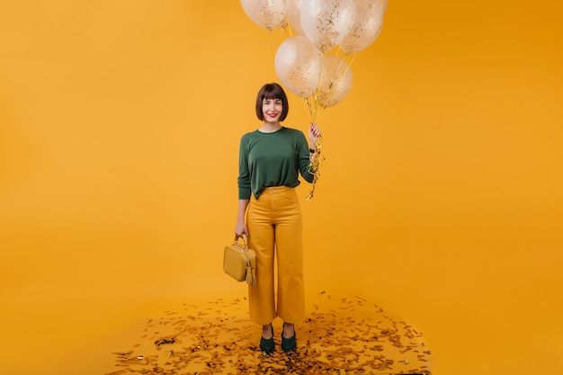 Retrato de cuerpo entero de una mujer bonita con bolso aislado. Filmación en interiores de elegante cumpleañera sosteniendo un montón de globos de fiesta.