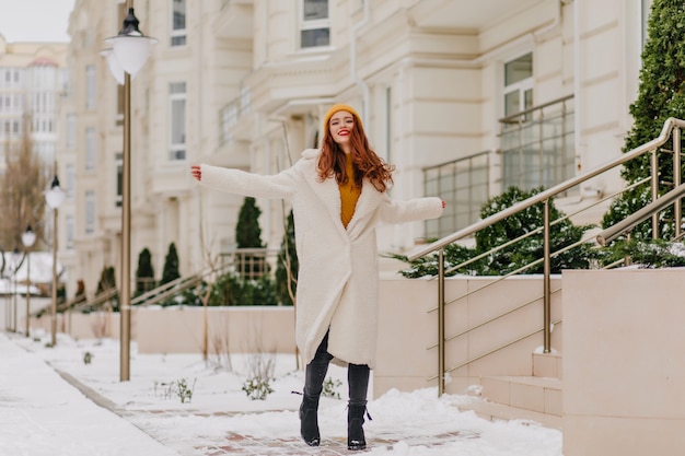 Retrato de cuerpo entero de una mujer caucásica alegre sonriendo en la calle nevada. Feliz chica de jengibre divirtiéndose en un día frío.