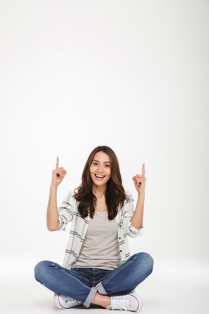 Foto gratuita retrato de cuerpo entero de mujer complacida en ropa casual sentado en postura de loto en el suelo y apuntando con los dedos índice hacia arriba, aislado sobre la pared blanca