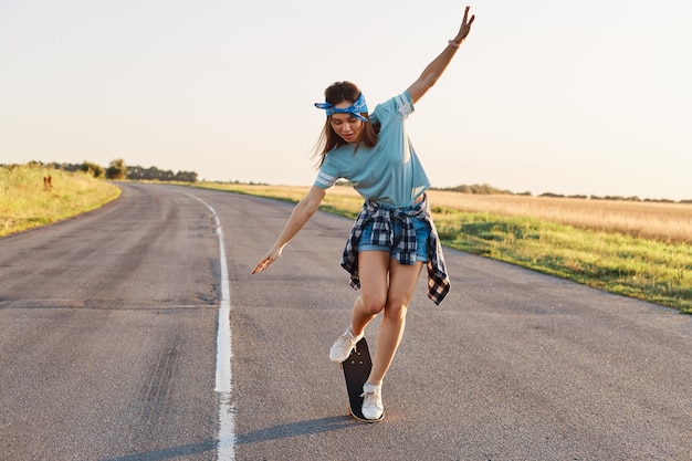 Retrato de cuerpo entero de una mujer deportiva delgada haciendo trucos en una patineta, pasando tiempo activo solo, al aire libre en la calle, brazos levantados, mirando hacia abajo con expresión facial emocionada.