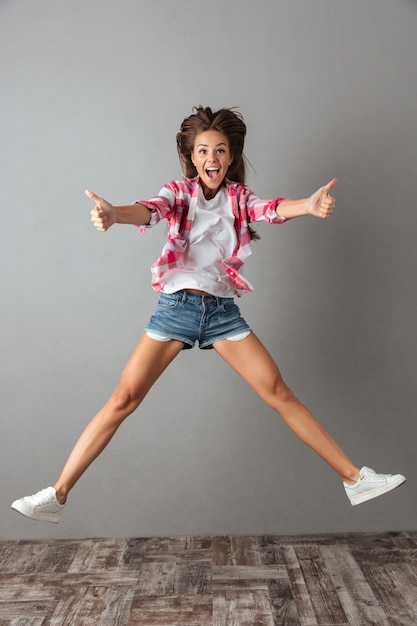 Foto gratuita retrato de cuerpo entero de mujer emocional saltando en ropa casual, mostrando el pulgar hacia arriba gesto