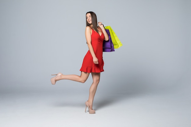 Retrato de cuerpo entero de una mujer feliz emocionada en vestido rojo de pie y sosteniendo coloridas bolsas de compras aisladas en una pared blanca