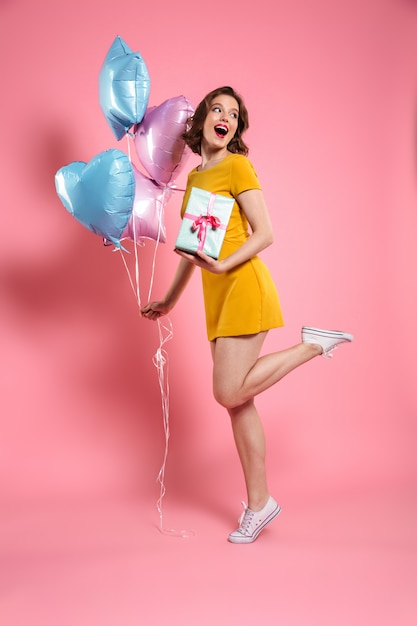 Retrato de cuerpo entero de una mujer joven feliz en vestido amarillo con caja de regalo y globos de colores, mirando a un lado