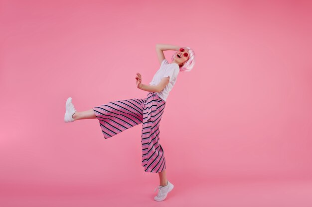 Retrato de cuerpo entero de una mujer joven en pantalones de moda bailando con una sonrisa feliz. Filmación en interiores de chica delgada y elegante con peluca rosa divirtiéndose