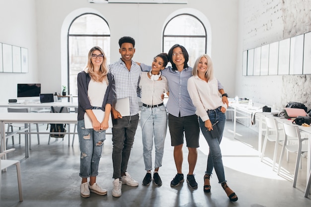 Retrato de cuerpo entero de una mujer rubia tímida en zapatillas blancas con portátil después del seminario y está al lado de un amigo africano. Estudiantes internacionales emocionados posando juntos después de la conferencia en un espacioso salón.