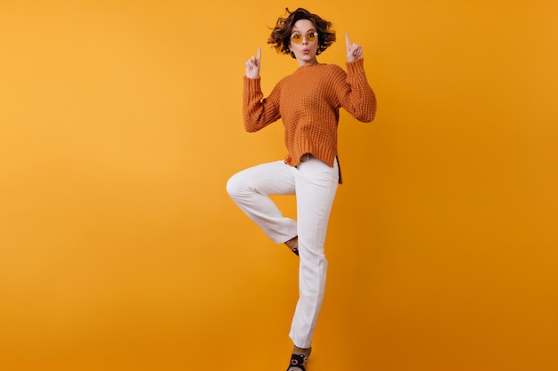 Retrato de cuerpo entero de una niña despreocupada en pantalones blancos saltando en el espacio naranja