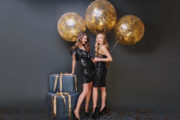 Retrato de cuerpo entero de una niña elegante con peinado de moda tocando la caja de regalo y riendo. dos damas extáticas posando con globos dorados.