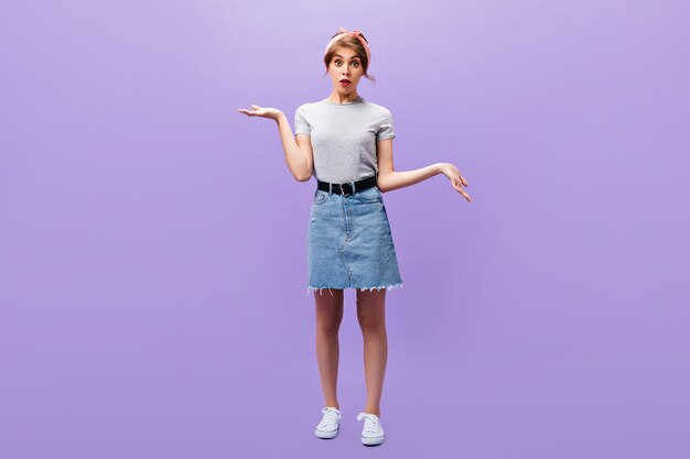 Retrato de cuerpo entero de niña en falda de mezclilla y camisa gris. Mujer sorprendida con diadema rosa en ropa elegante y zapatillas blancas posando.