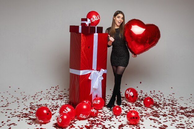 Retrato de cuerpo entero de una niña morena de raza blanca con un globo en forma de corazón apuntando con su pulgar a los regalos envueltos. Globos de aire con cartel de venta y descuento en el piso con confeti.