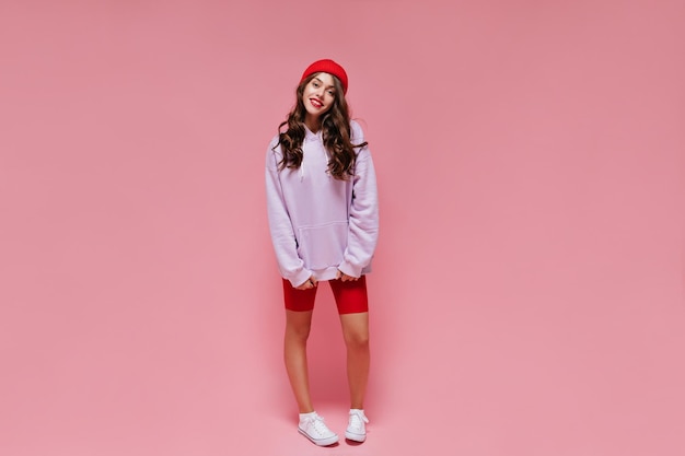 Foto gratuita retrato de cuerpo entero de niña morena rizada en shorts de ciclismo rojos y sudadera con capucha púrpura sobre fondo rosa aislado mujer feliz en poses de sombrero brillante y sonrisas