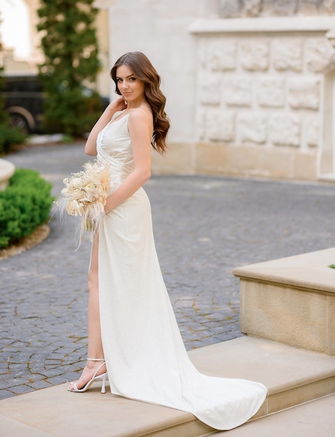 Retrato de cuerpo entero de la novia elegante morena en vestido de novia largo de seda con ramo de flores Hermosa mujer joven posando al aire libre en vestido con hombros abiertos Peinado de boda