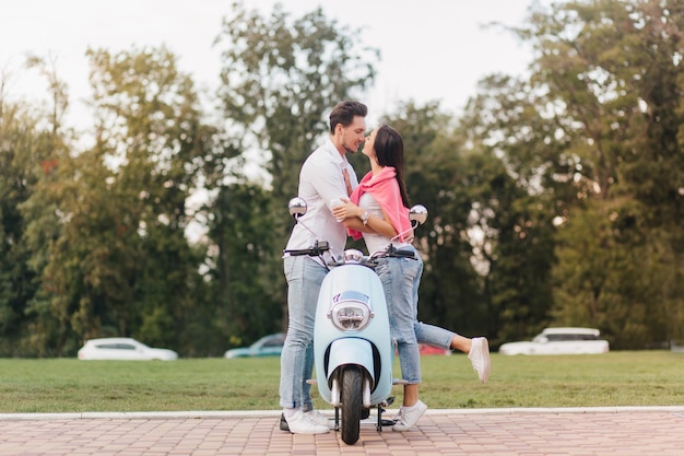 Foto gratuita retrato de cuerpo entero de la pareja besándose posando cerca de scooter con árboles en el fondo. agraciada chica de pie sobre una pierna y cogidos de la mano con su novio con camisa blanca en la mañana en el parque.