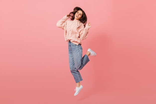 Retrato de cuerpo entero de saltar niña morena. Señora en jeans y suéter rosa divirtiéndose sobre fondo aislado.