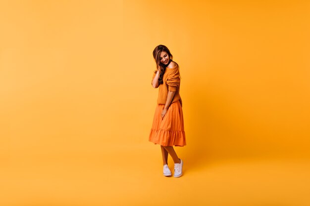 Retrato de cuerpo entero de una tímida niña caucásica de pie en amarillo. Filmación en interiores de una chica encantadora con falda y jersey de punto naranja.