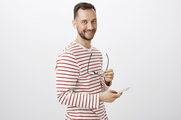 Retrato de curioso guapo gay europeo en jersey de rayas, quitándose las gafas y esperando la llamada de su novio en auriculares inalámbricos