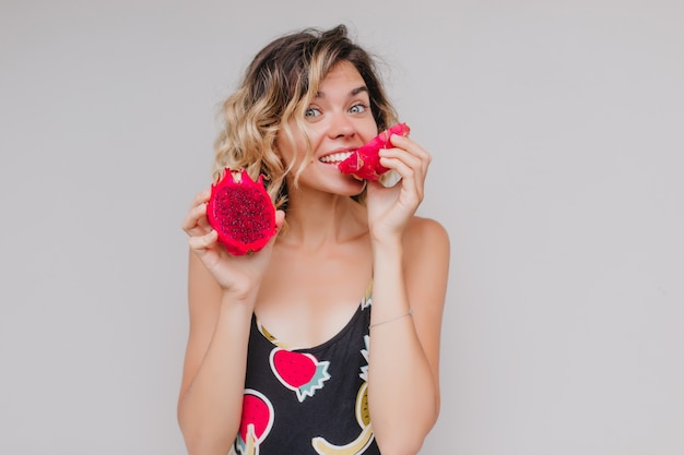 retrato de dama alegre con piel bronceada posando con sabrosa pitahaya. Foto interior de hermosa niña con peinado rizado sosteniendo fruta del dragón y sonriendo.