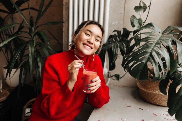 Retrato de dama alegre en suéter rojo de gran tamaño sosteniendo un vaso de jugo de sandía recién exprimido, sentado en el café contra la pared de las plantas