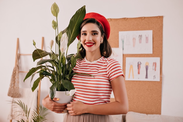 Retrato de dama en camisa a rayas tiene planta. Mujer bonita en camiseta ligera y boina roja posando en cámara con flor en sus manos.