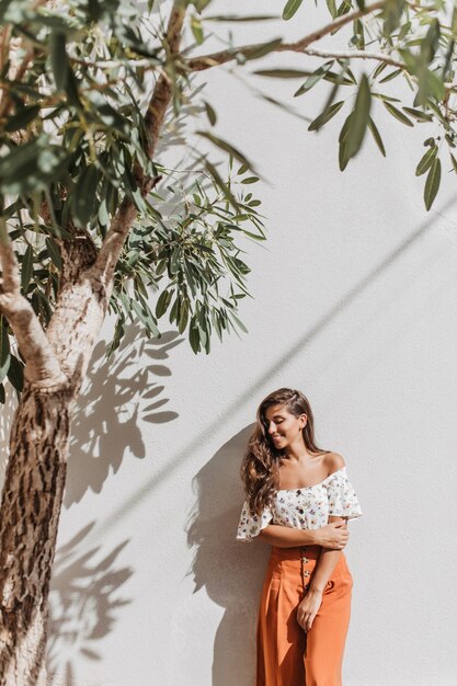 Retrato de dama encantadora en traje de resort de verano posando junto al olivo en la pared blanca