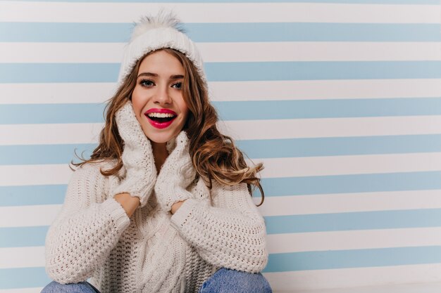 Retrato de dama entusiasta y emocional en ropa cómoda y suave y accesorios de invierno sentado en el piso contra la pared azul-blanca