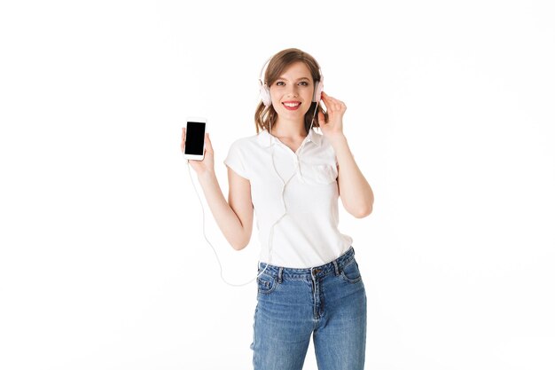 Retrato de una dama muy sonriente con auriculares de pie con el celular en la mano y felizmente mirando a la cámara sobre fondo blanco aislado