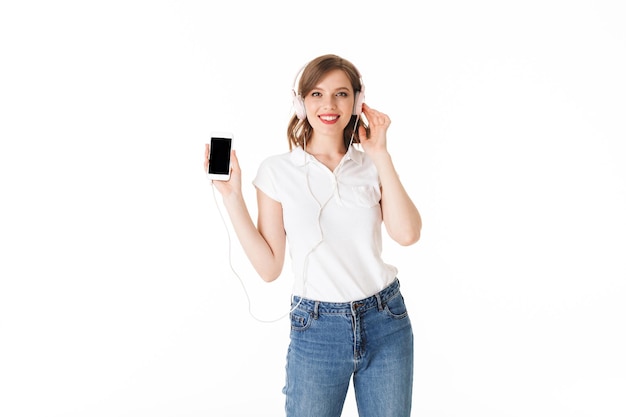 Retrato de una dama muy sonriente con auriculares de pie con el celular en la mano y felizmente mirando a la cámara sobre fondo blanco aislado