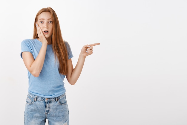 Retrato de dama pelirroja caucásica sorprendida y sorprendida con pecas jadeando abriendo la boca tocando la mejilla y apuntando a la derecha siendo interrogada y curiosa mientras presencia una escena impactante