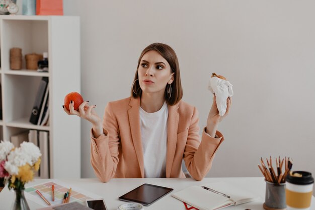 Retrato de dama con pelo corto en la oficina. Mujer de negocios en pensamiento elige comer hamburguesa o manzana saludable.