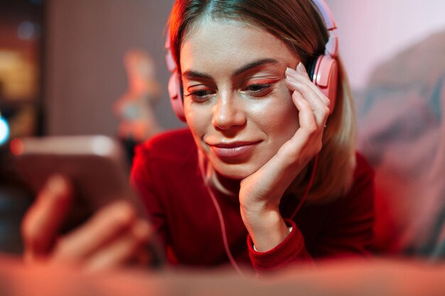 Retrato de dama pensativa en auriculares acostado en el sofá con teléfono celular en las manos mientras escucha música en casa